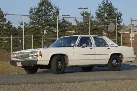 1991 Ford Crown Victoria LTD - A white sedan is parked outdoors next to a chain-link fence with trees in the background.