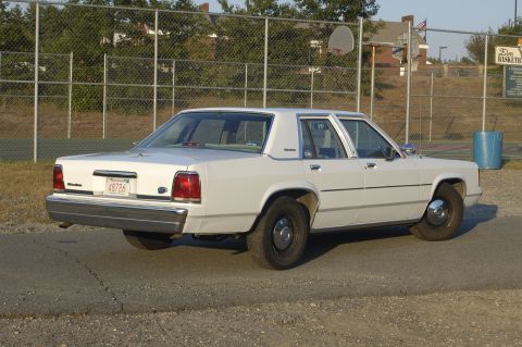 1991 Ford Crown Victoria LTD - A white sedan is parked on a road with a chain-link fence and basketball court in the background.