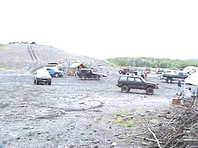 Several off-road vehicles are parked in a gravel lot with tents and trees in the background