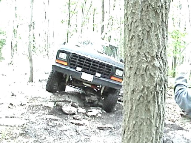 Dark Ford Bronco driving over rocks, front grille and headlights visible