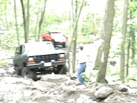 1978 Ford Bronco - A 1978 Ford Bronco navigates a rocky trail in a wooded area, with a red vehicle visible in the background.