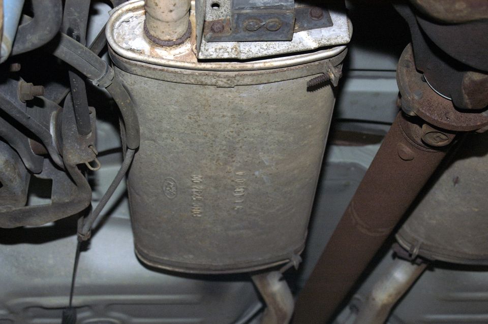 Close-up of a car's muffler with part numbers visible. Driveshaft and other exhaust components are also in the shot
