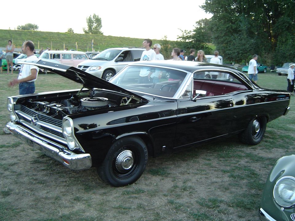Black 1960s Ford Fairlane hardtop with hood up showing V8, dog-dish caps, at grass car show