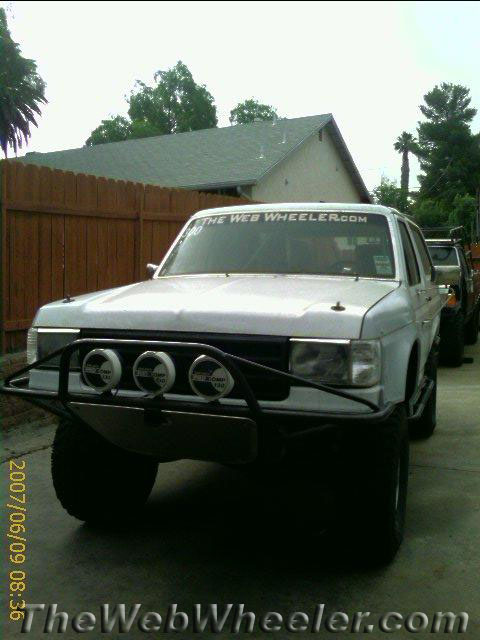 1990 Ford Bronco - white lifted SUV with a tubular front bumper and three round driving lights parked in a fenced backyard driveway.