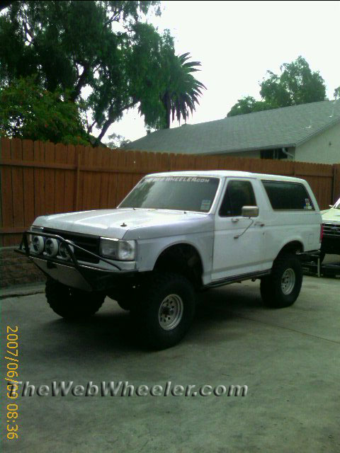 1990 Ford Bronco - white lifted Ford Bronco on large off-road tires with a tube bumper and driving lights, parked on a concrete pad.