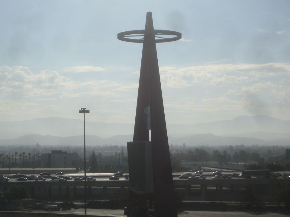 Tall steel spire with ring tower silhouetted against hazy sky over a parking lot
