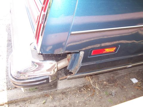 1999 Ford Crown Victoria - The rear of a blue car shows a chrome bumper, exhaust pipe, and a damaged black trim piece.
