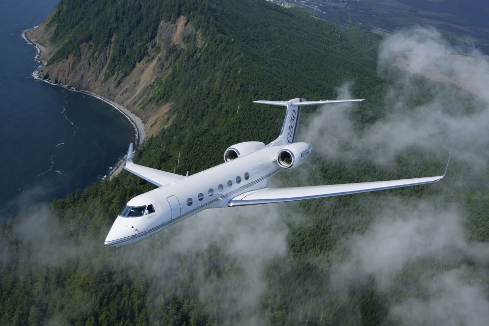 White private jet flying over a forested coastline, with clouds obscuring the land below