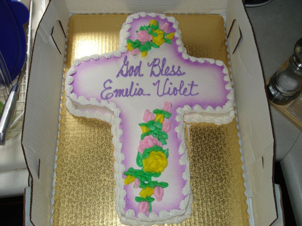 cross-shaped cake with purple and white frosting, reads 'God Bless Emilia Violet', decorated with pink and yellow flowers