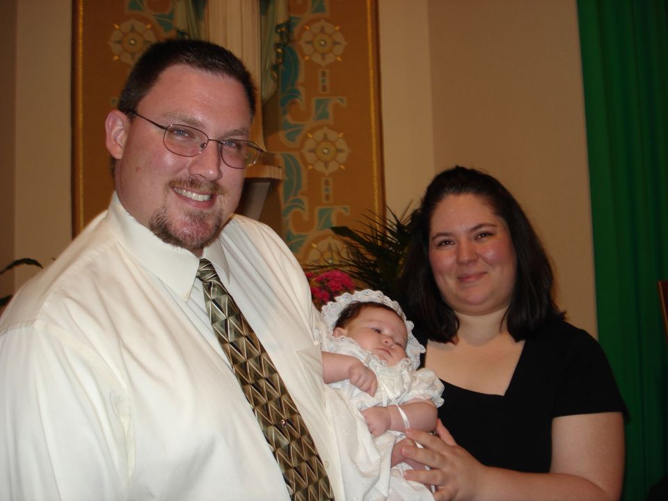 Man in a white shirt and tie holds a baby in a christening gown, with a woman behind them