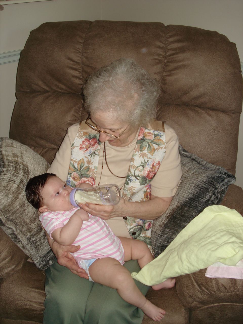 Elderly woman with glasses feeding a baby with a bottle while sitting in a recliner