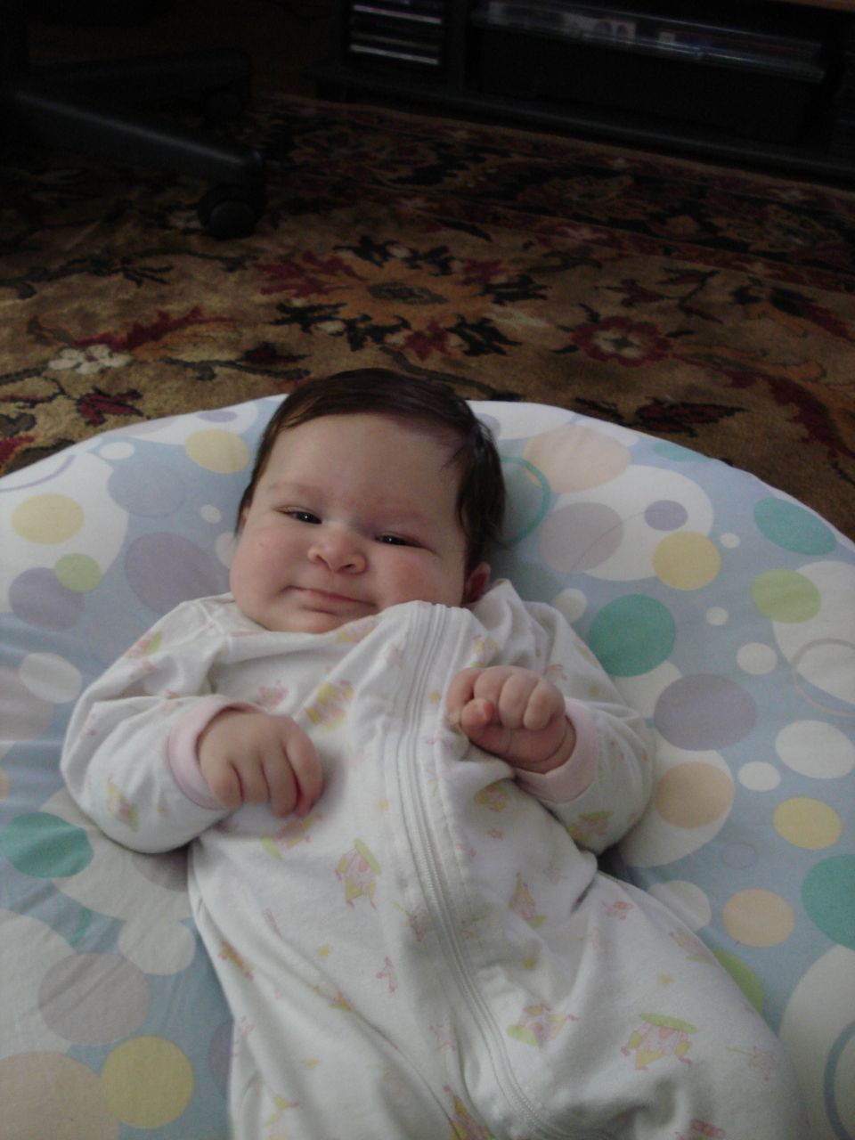 Baby lying on a patterned pillow, wearing a white zippered onesie with cartoon prints