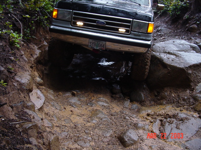 Front of a dark Ford truck flexed over rocks in a muddy rutted off-road trail
