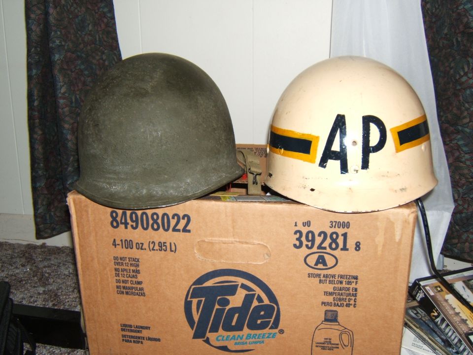 Two military helmets, one olive drab and one cream with black and yellow bands and 'AP' marking, sit atop a cardboard box