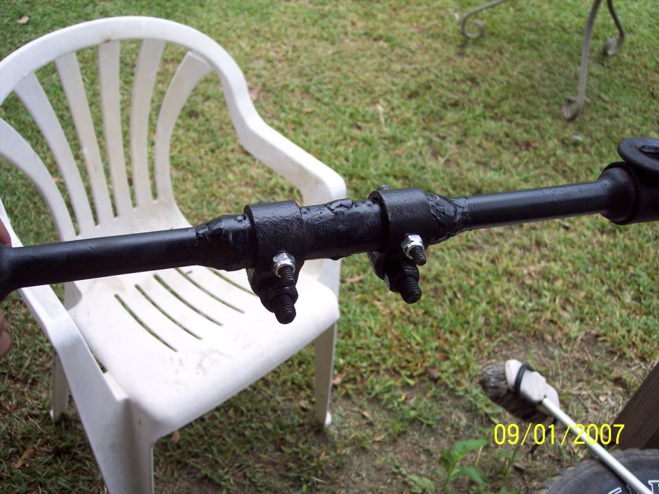 Black metal steering linkage component with two clamps and bolts, resting on a white plastic chair