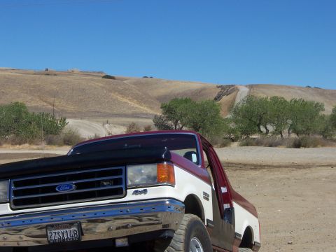 1991 Ford Bronco - A two-tone Ford truck is parked on a dirt road with rolling hills and trees under a clear blue sky.