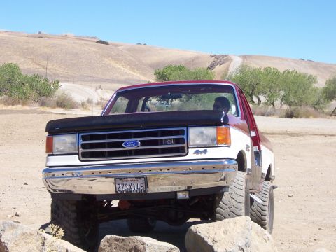 1991 Ford Bronco - A red and white Ford F-Series truck is shown from the front, navigating over rocks on a dry, hilly landscape.