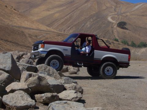 1991 Ford Bronco - A man drives a red and white Ford Bronco over a rocky terrain with hills in the background.