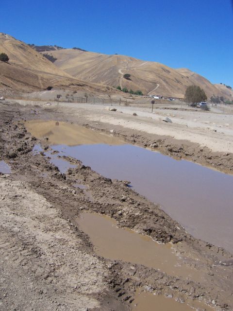 1991 Ford Bronco - A muddy track with several large puddles, set against a backdrop of dry, rolling hills under a clear blue sky.