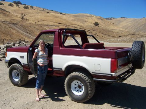1991 Ford Bronco - A woman stands next to a two-tone Ford Bronco in a dry, hilly landscape.