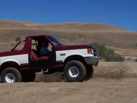 1991 Ford Bronco - A driver maneuvers a two-tone Ford Bronco through a dusty, off-road terrain under a clear blue sky.