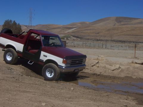 1991 Ford Bronco - A two-tone Ford Bronco navigates a muddy track in a dry, hilly landscape under a clear blue sky.
