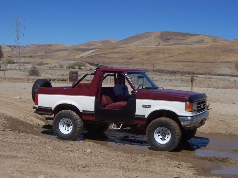 1991 Ford Bronco - A maroon and white 1991 Ford Bronco is shown off-road, with its doors removed and a spare tire mounted at the rear.