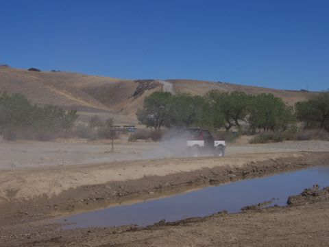 1991 Ford Bronco - A two-tone pickup truck drives through a dusty, arid landscape with trees and hills in the background.