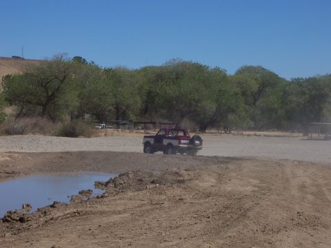 1991 Ford Bronco - A red and white Ford Bronco drives on a dusty dirt road near a muddy puddle.