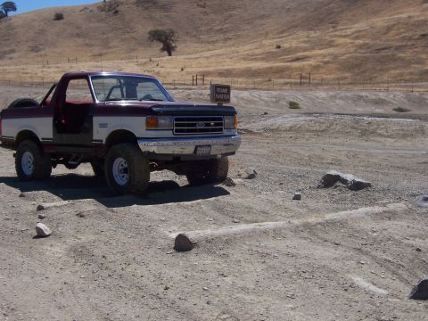1991 Ford Bronco - A two-tone Ford Bronco sits on a dusty trail with a sign reading 'Frame Twister' in the background.