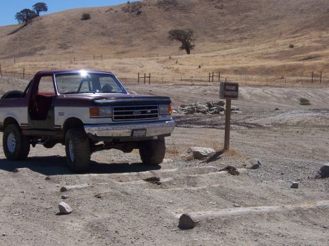 1991 Ford Bronco - A maroon and white 1991 Ford Bronco with off-road tires sits parked on a dirt track near a sign that reads "Frame Twister."