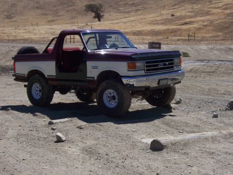 1991 Ford Bronco - A two-tone Ford Bronco with large tires sits on a dirt lot, its driver's door ajar.