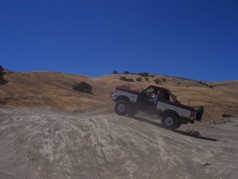 1991 Ford Bronco - A two-tone Ford Bronco navigates an off-road trail under a clear blue sky.