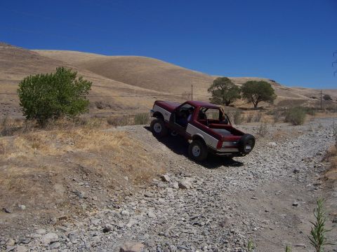1991 Ford Bronco - A red and silver SUV navigates a rocky, off-road path under a clear blue sky.