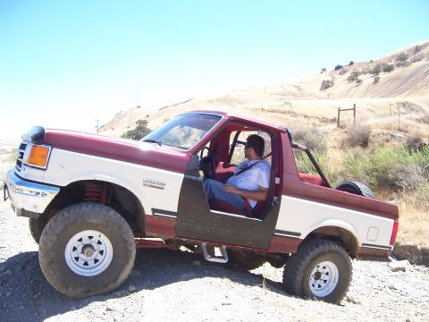 1991 Ford Bronco - A man sits in a lifted Ford Bronco with its doors off, parked on a dusty, off-road trail under a clear blue sky.