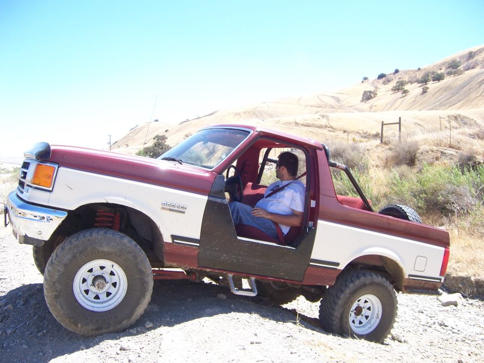 Man in a red and white Ford Bronco with doors removed, parked on a dirt road
