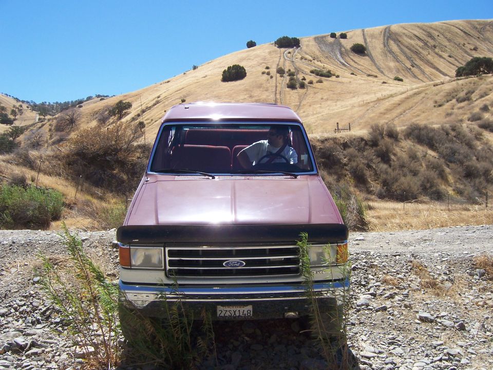 Maroon Ford truck with a black bug deflector, parked on a dirt road. A man is driving