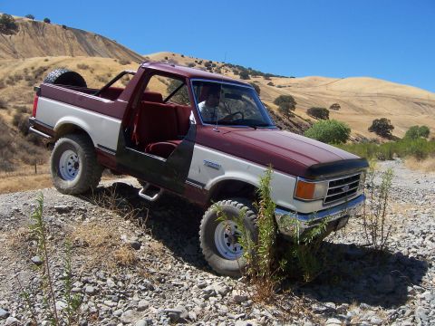 1991 Ford Bronco - A maroon and white 1991 Ford Bronco with its doors removed navigates a rocky, off-road trail under a clear blue sky.