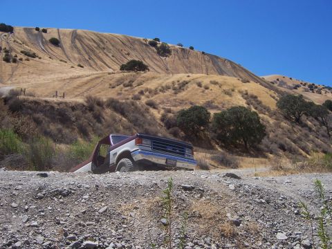 1991 Ford Bronco - A two-tone Ford F-Series truck is partially stuck on a rocky, dirt path with a large, dry hill behind it.