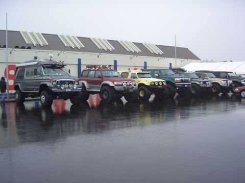 1996 Ford Bronco - A line of modified off-road vehicles are parked outside a building on a wet day.
