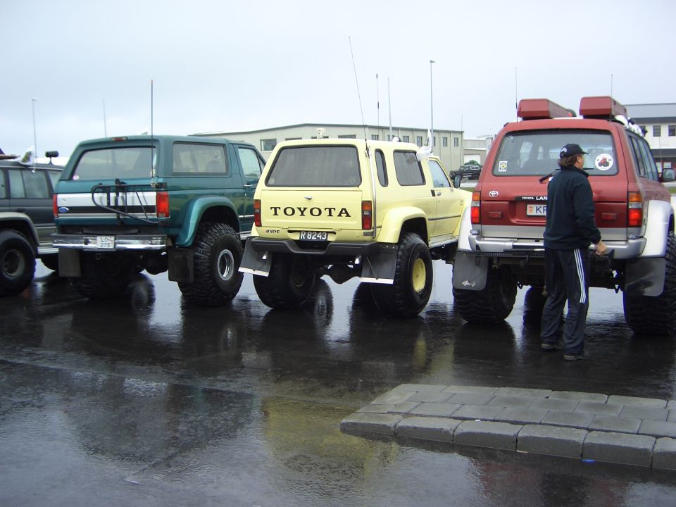 Three lifted 4x4 SUVs parked rear-facing on wet lot, teal Ford Bronco, yellow Toyota, red Land Cruiser