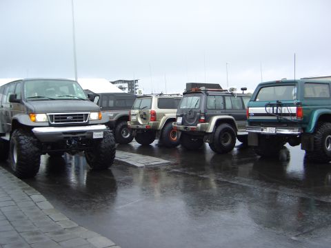 1996 Ford Bronco - A collection of off-road vehicles, including a van and several SUVs, are parked together on a wet, paved surface.