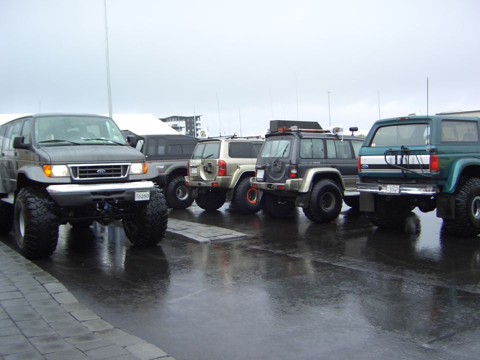 Row of lifted 4x4 SUVs and a van on big tires on wet lot, green Ford Bronco at right