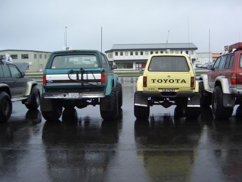 1996 Ford Bronco - Several modified off-road vehicles are parked on a wet surface, viewed from the rear.