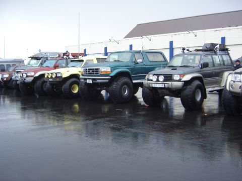 1996 Ford Bronco - Several modified SUVs, including a green 1996 Ford Bronco, are lined up on a wet asphalt surface.