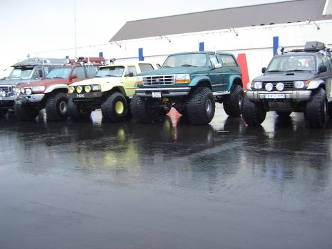 1996 Ford Bronco - A lineup of modified SUVs with large tires sits on a wet asphalt surface in front of a building.