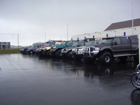 1996 Ford Bronco - A row of modified trucks and SUVs are parked on a wet asphalt lot under an overcast sky.