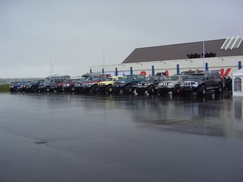 1996 Ford Bronco - Multiple SUVs and vans are parked in a line in front of a large building under a cloudy sky.