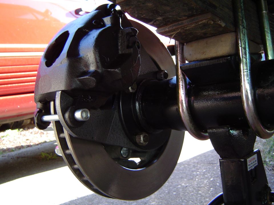 Black brake caliper and rotor assembly on a vehicle axle, with a red vehicle body visible in the background