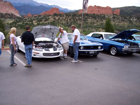 1972 Ford Ranchero - Several cars are parked in a lot with people looking at them, set against a backdrop of mountains and red rock formations.